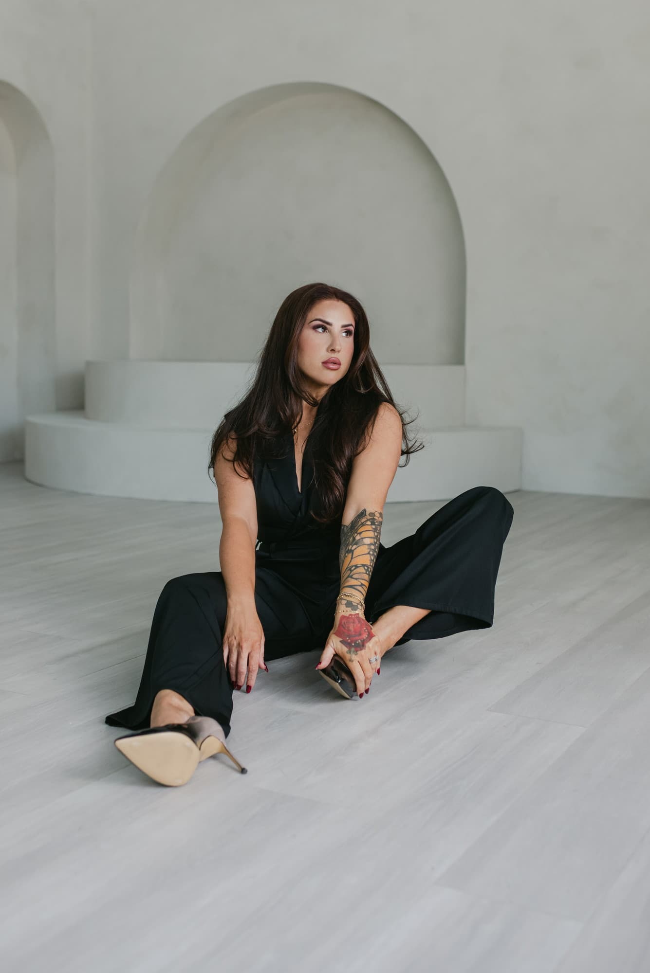 Meagan Greene seated on a studio floor in a black jumpsuit and heels